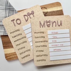 May include: Two light-colored wooden boards with rounded corners, featuring a repeating mouse-ear pattern. One board is labeled "TO DO" and the other "MENU," both with days of the week listed. The menu board has white strips with handwritten food items.