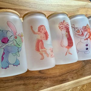 K&ouml;nnte beinhalten: F&uuml;nf mattierte Gl&auml;ser mit Bambusdeckeln, jedes mit einer anderen farbenfrohen Zeichentrickfigur-Illustration. Die Motive zeigen Stitch mit einer Puppe, Moana, Cruella de Vil, Olaf mit Sven sowie Minnie Maus in einem blauen Outfit.