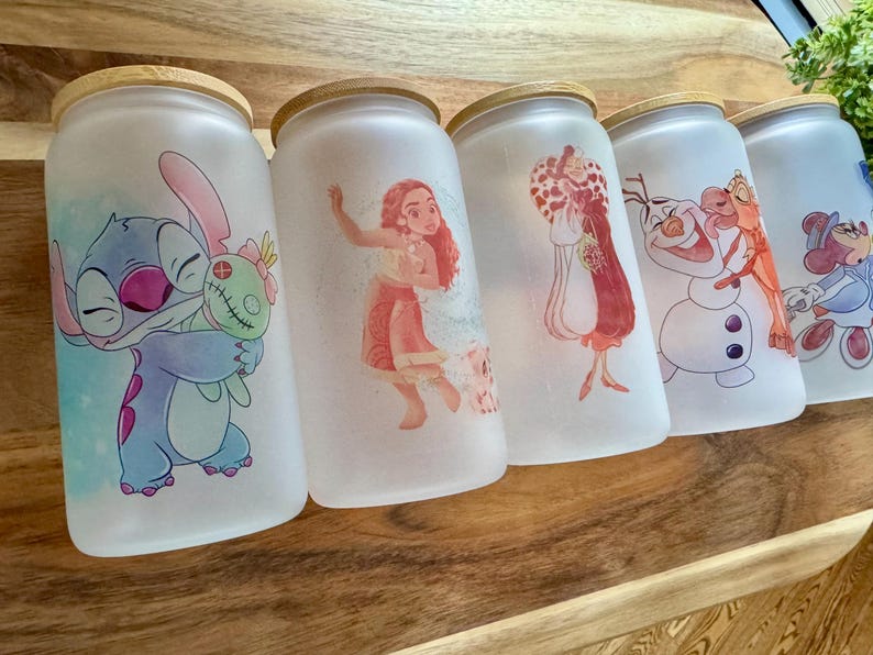 K&ouml;nnte beinhalten: F&uuml;nf mattierte Gl&auml;ser mit Bambusdeckeln, jedes mit einer anderen farbenfrohen Zeichentrickfigur-Illustration. Die Motive zeigen Stitch mit einer Puppe, Moana, Cruella de Vil, Olaf mit Sven sowie Minnie Maus in einem blauen Outfit.