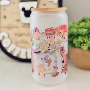 K&ouml;nnte beinhalten: Ein mattiertes Glas mit Bambusdeckel, das mit einem farbenfrohen Grafikdruck von Zeichentrickfiguren wie einem Weltraum-Ranger, einem Cowboy, einem Cowgirl und einem Hund versehen ist. Das Design umschlie&szlig;t die zylindrische Glasoberfl&auml;che.