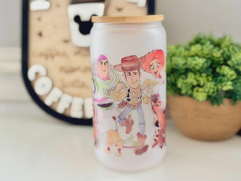 K&ouml;nnte beinhalten: Ein mattiertes Glas mit Bambusdeckel, das mit einem farbenfrohen Grafikdruck von Zeichentrickfiguren wie einem Weltraum-Ranger, einem Cowboy, einem Cowgirl und einem Hund versehen ist. Das Design umschlie&szlig;t die zylindrische Glasoberfl&auml;che.