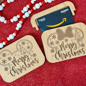 May include: Wooden Christmas gift card holders with a Disney theme. One holder contains an Amazon gift card. The holders are engraved with snowflakes, stars, a Christmas tree, and the words "Merry Christmas". A red and white beaded garland is in the background.