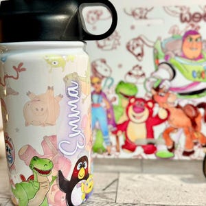 Puede incluir: Una botella de agua blanca con personajes de Toy Story, como Buzz Lightyear y Woody. El nombre "Emma" est&aacute; escrito verticalmente en cursiva. La botella tiene una tapa negra. El fondo es blanco.