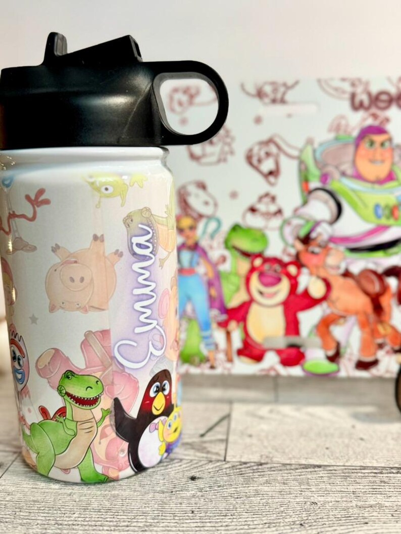 Puede incluir: Una botella de agua blanca con personajes de Toy Story, como Buzz Lightyear y Woody. El nombre "Emma" est&aacute; escrito verticalmente en cursiva. La botella tiene una tapa negra. El fondo es blanco.