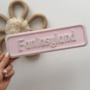 Puede incluir: Un letrero rectangular rosa claro con la palabra "Fantasyland" en letras blancas en relieve. El letrero tiene un borde blanco. Una decoración beige en forma de flor está en el fondo.