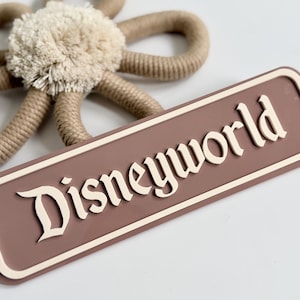 Puede incluir: Un letrero rectangular con la palabra "Disneyworld" en una fuente estilizada. El letrero es de color malva apagado con un borde y letras de color crema. Una flor decorativa hecha de cuerda y un centro esponjoso en el fondo.