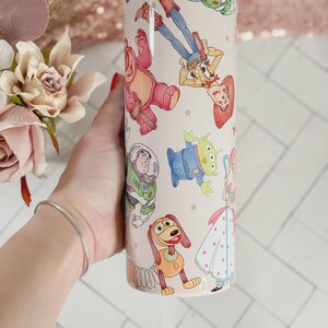 May include: A cylindrical white tumbler featuring a colorful, repeating pattern of various animated characters, including a cowboy, a space ranger, a green alien, a pink bear, a spring-based dog, and a shepherdess, all set against a light background.