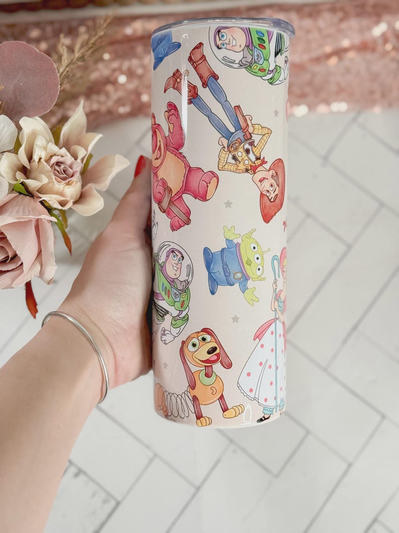 May include: A cylindrical white tumbler featuring a colorful, repeating pattern of various animated characters, including a cowboy, a space ranger, a green alien, a pink bear, a spring-based dog, and a shepherdess, all set against a light background.