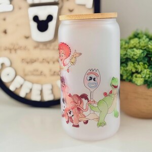 K&ouml;nnte beinhalten: Ein mattierter Glastrinkbecher mit Bambusdeckel, der mit einem farbenfrohen Rundumdruck von Zeichentrickfiguren versehen ist, darunter eine Cowgirl-Figur, eine gabelartige Figur mit Wackelaugen und ein gr&uuml;ner Dinosaurier.