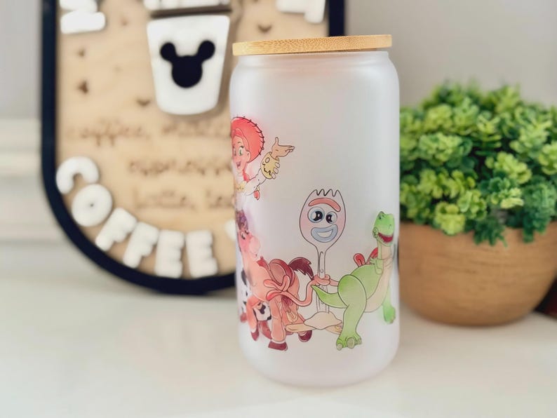 K&ouml;nnte beinhalten: Ein mattierter Glastrinkbecher mit Bambusdeckel, der mit einem farbenfrohen Rundumdruck von Zeichentrickfiguren versehen ist, darunter eine Cowgirl-Figur, eine gabelartige Figur mit Wackelaugen und ein gr&uuml;ner Dinosaurier.