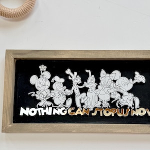 May include: A framed artwork featuring Disney characters in a line, with the phrase "NOTHING CAN STOP US NOW" in gold lettering. The characters are outlined in white against a black background, within a wooden frame.