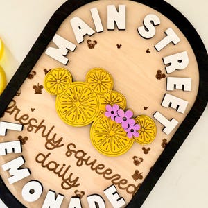 May include: An oval wooden sign with a black border and light wood background. It features white 3D lettering that reads MAIN STREET and LEMONADE, with the text freshly squeezed daily in brown. A yellow lemon-slice mouse-shaped icon has purple flowers.