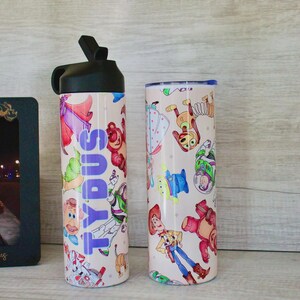 May include: A black plastic water bottle with a straw lid and a cylindrical tumbler, both featuring a light-colored surface decorated with colorful cartoon character illustrations. The water bottle displays the name TYDUS in vertical purple letters.