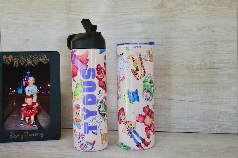 May include: A black plastic water bottle with a straw lid and a cylindrical tumbler, both featuring a light-colored surface decorated with colorful cartoon character illustrations. The water bottle displays the name TYDUS in vertical purple letters.