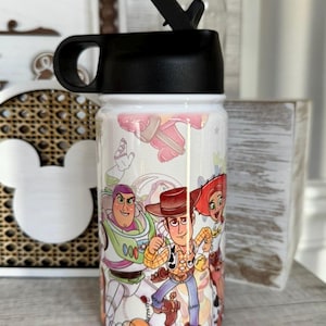 Puede incluir: Una botella de agua blanca con tapa negra y boquilla, decorada con personajes de "Toy Story". El dise&ntilde;o incluye a Woody, Buzz Lightyear y Jessie. Adecuada para llevar agua u otras bebidas.