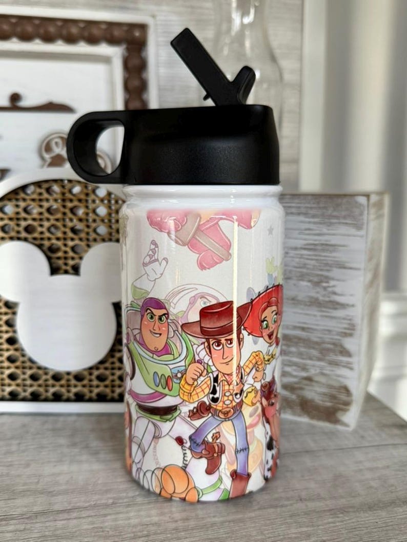 Puede incluir: Una botella de agua blanca con tapa negra y boquilla, decorada con personajes de "Toy Story". El dise&ntilde;o incluye a Woody, Buzz Lightyear y Jessie. Adecuada para llevar agua u otras bebidas.