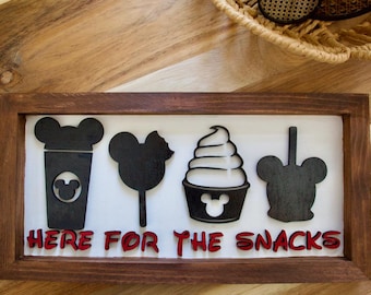 Könnte beinhalten: Holzgerahmtes Schild mit schwarzen Ausschnitten eines Getränks, eines Lollipops, eines Eis und eines Mickey Mouse-Kopfes. Der rote Text unten lautet "HERE FOR THE SNACKS". Das Schild befindet sich auf einer Holzoberfläche.