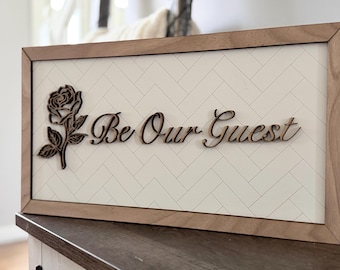 May include: Wooden framed sign with the words "Be Our Guest" in a cursive font. A rose with leaves is on the left side. The background is white with a herringbone pattern. The frame is a light brown color.