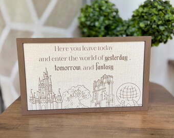 May include: A rectangular wooden sign with a light beige fabric center featuring the brown text: Here you leave today and enter the world of yesterday, tomorrow, and fantasy. Below the text are brown line art illustrations of a castle, a tree, a tower, and a globe.