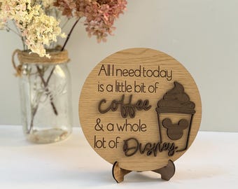 Könnte beinhalten: Runder Holzschild mit dem Text "All I need today is a little bit of coffee & a whole lot of Disney". Das Schild zeigt eine Kaffeetasse mit einem Mickey Mouse-Kopf-Design. Das Schild steht auf einem Holzständer.