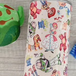 May include: A cylindrical tumbler featuring a repeating pattern of various animated characters including a space ranger, a cowgirl, a shepherdess, a spring-based dog, and an alien. The background is a light cream color with small gray star accents.