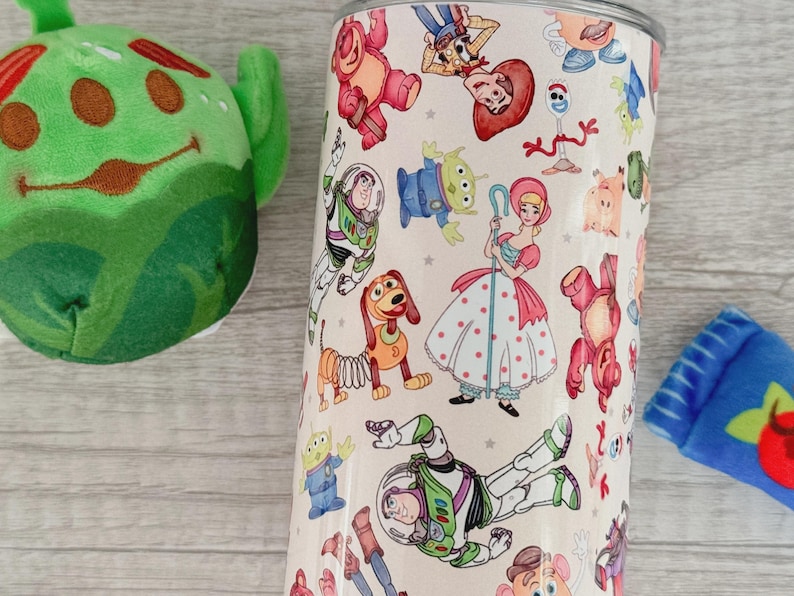 May include: A cylindrical tumbler featuring a repeating pattern of various animated characters including a space ranger, a cowgirl, a shepherdess, a spring-based dog, and an alien. The background is a light cream color with small gray star accents.