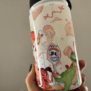 Puede incluir: Una botella de agua blanca con tapa negra, decorada con ilustraciones coloridas de personajes de la pel&iacute;cula animada Toy Story. La botella presenta im&aacute;genes de Woody, Buzz Lightyear y otros personajes.