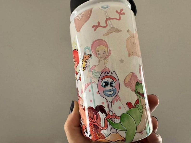 Puede incluir: Una botella de agua blanca con tapa negra, decorada con ilustraciones coloridas de personajes de la pel&iacute;cula animada Toy Story. La botella presenta im&aacute;genes de Woody, Buzz Lightyear y otros personajes.
