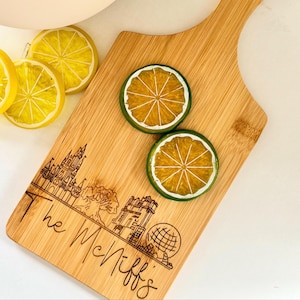 May include: A bamboo cutting board with a handle, featuring a laser-etched design of a castle and other structures, with the name "The McNiff's" below. Two lime slice coasters and lemon slices are on the board.
