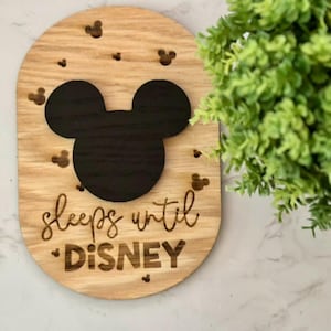 May include: Wooden sign with a light brown finish, featuring a black Mickey Mouse head silhouette and smaller Mickey Mouse shapes. The sign reads "sleeps until DISNEY" in a script font. A green plant is in the background.