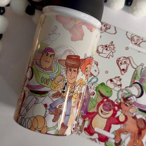 Puede incluir: Una botella de agua blanca con ilustraciones coloridas de personajes de la pel&iacute;cula animada "Toy Story". La botella tiene una tapa negra y un dise&ntilde;o a juego en una tarjeta separada. Los personajes incluyen a Buzz Lightyear y Woody.