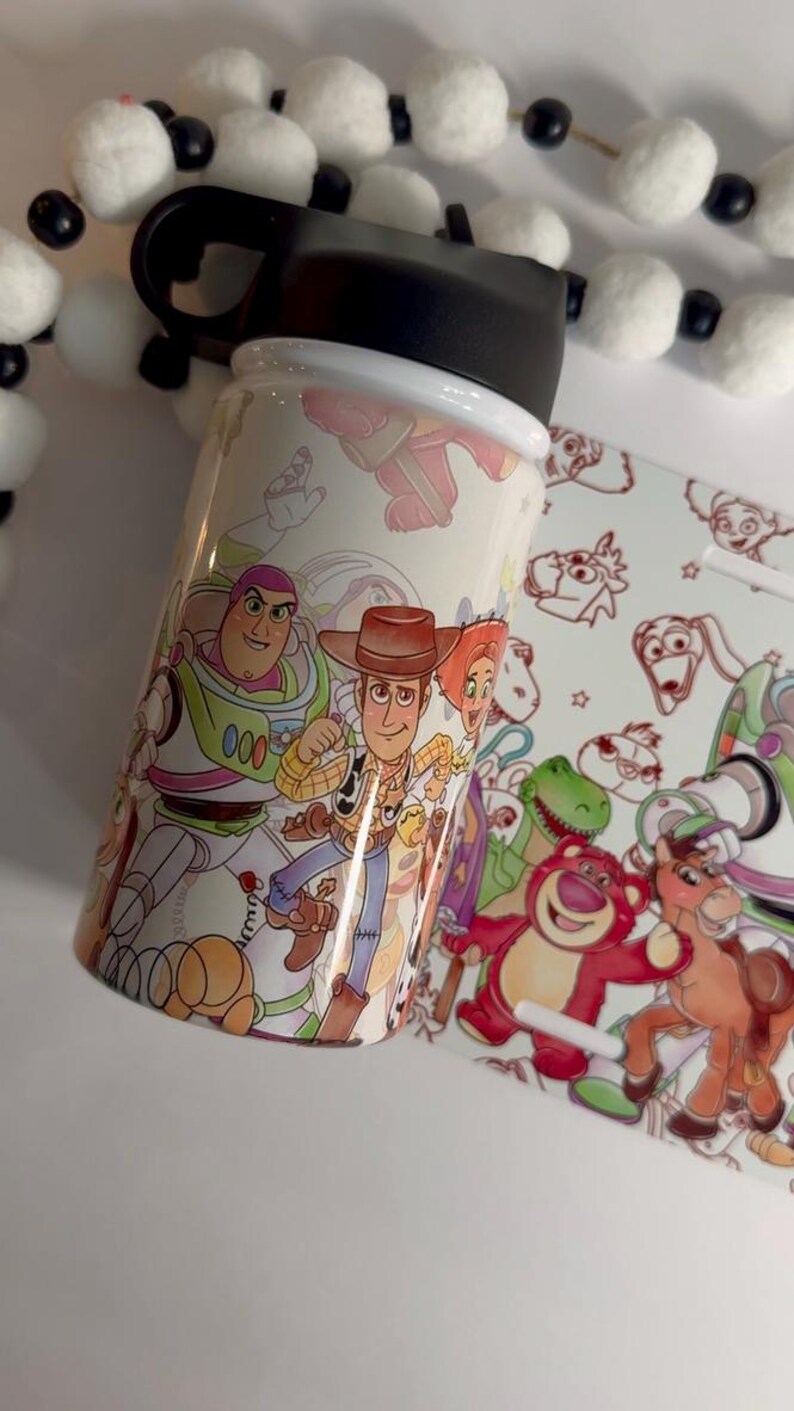 Puede incluir: Una botella de agua blanca con ilustraciones coloridas de personajes de la pel&iacute;cula animada "Toy Story". La botella tiene una tapa negra y un dise&ntilde;o a juego en una tarjeta separada. Los personajes incluyen a Buzz Lightyear y Woody.