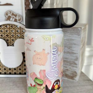 Puede incluir: Una botella de agua blanca con tapa y asa negras, con un dise&ntilde;o de dibujos animados colorido. La botella est&aacute; decorada con varios personajes de dibujos animados y el nombre "Emma".