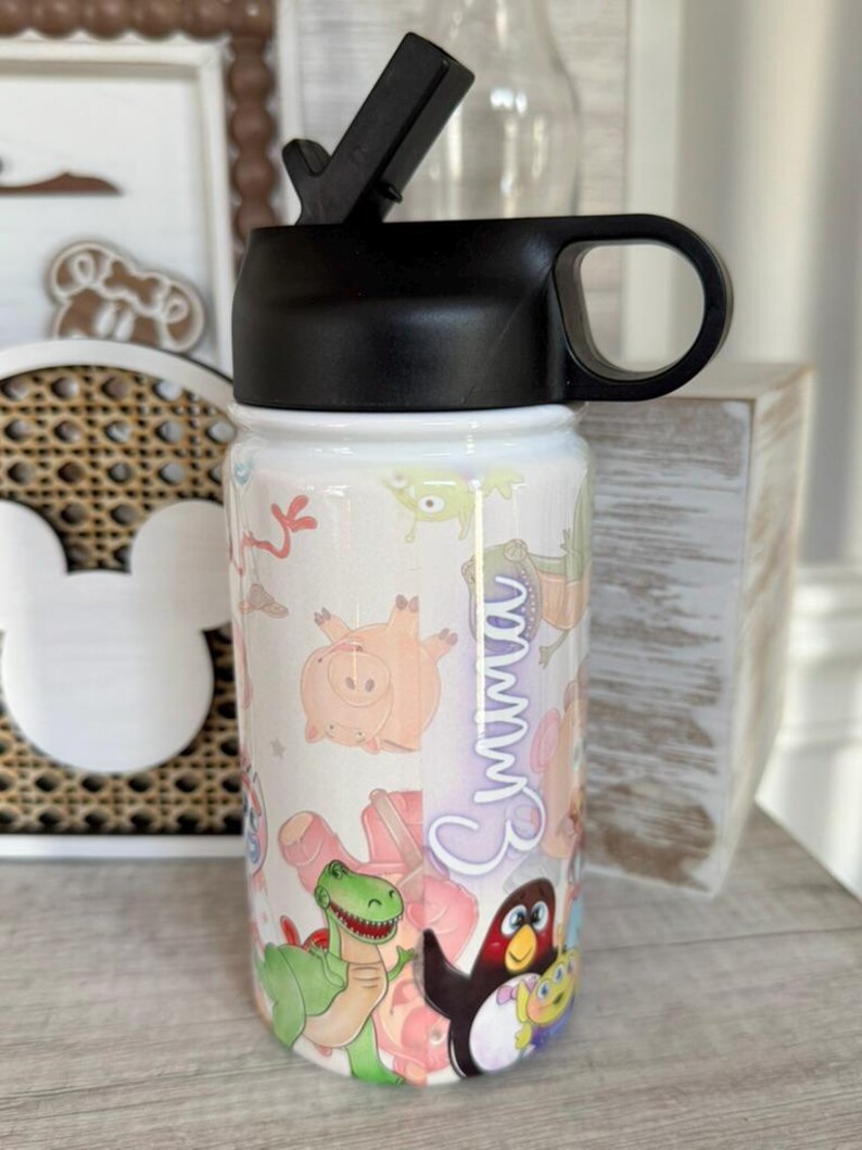 Puede incluir: Una botella de agua blanca con tapa y asa negras, con un dise&ntilde;o de dibujos animados colorido. La botella est&aacute; decorada con varios personajes de dibujos animados y el nombre "Emma".