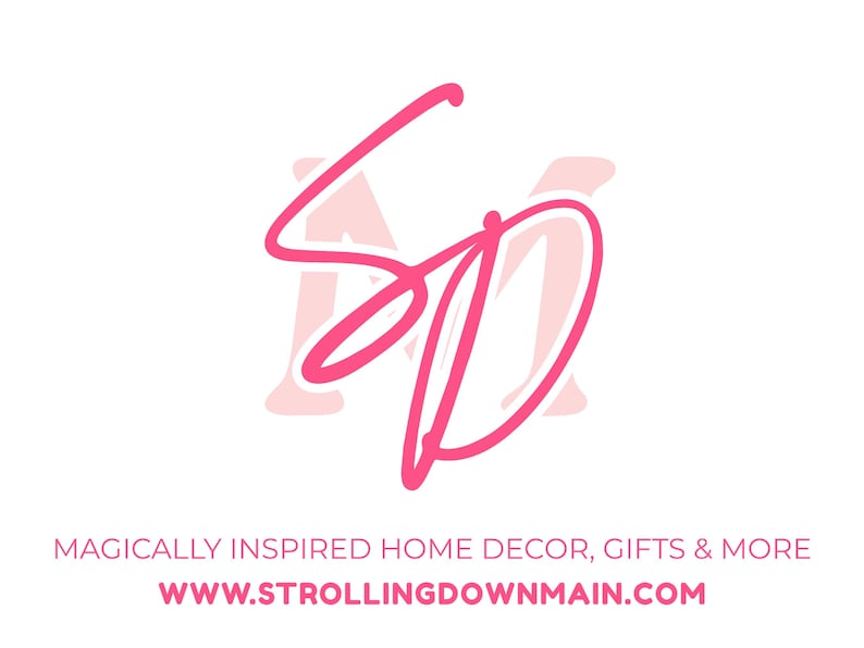 May include: A logo featuring the pink script letters SD overlaid on a pale pink letter M. Below the logo, the text reads MAGICALLY INSPIRED HOME DECOR, GIFTS & MORE followed by the website address WWW.STROLLINGDOWNMAIN.COM in pink capital letters.