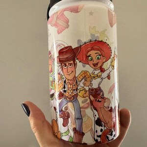 Puede incluir: Un vaso blanco con personajes de dibujos animados coloridos Woody, Jessie y Bullseye de la pel&iacute;cula Toy Story. El vaso tiene una tapa negra y lo sostiene una mano.