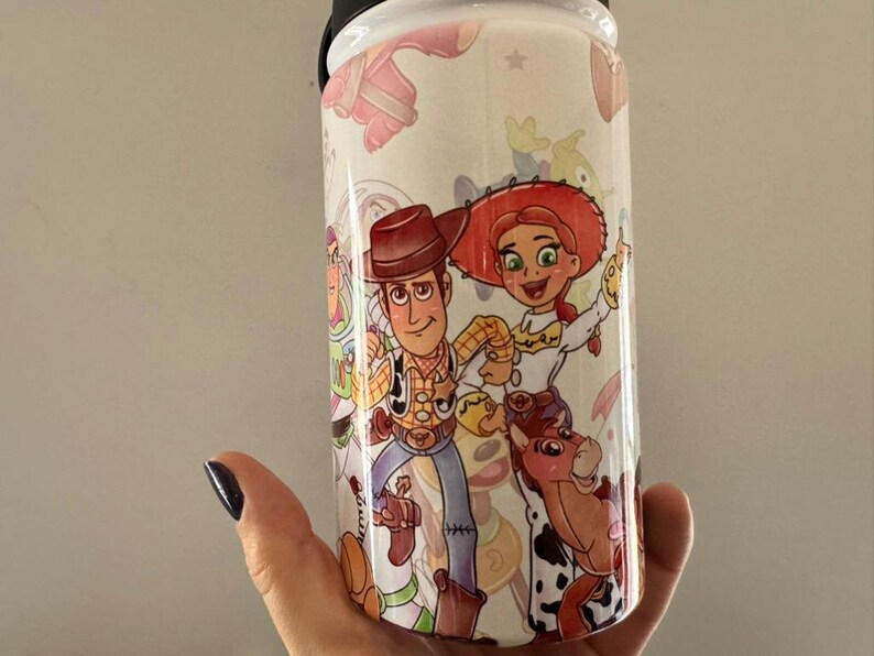 Puede incluir: Un vaso blanco con personajes de dibujos animados coloridos Woody, Jessie y Bullseye de la pel&iacute;cula Toy Story. El vaso tiene una tapa negra y lo sostiene una mano.