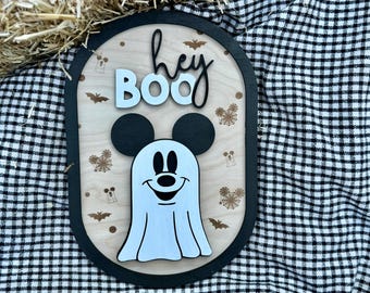 May include: An oval wooden wall sign with a black frame featuring the text "hey BOO" in black and white lettering. Below the text is a layered wooden cutout of a white ghost with black mouse ears, eyes, and nose. The background has small brown icons.