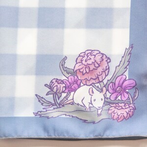 May include: A blue and white gingham fabric square with a white mouse sitting on a bed of pink and purple flowers. The fabric has a blue border.