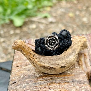 May include: A black macramé ring featuring a silver-coloured rose detail and a black bead. The ring is presented on a piece of driftwood, emphasising its handcrafted nature. The blurred background draws attention to the ring's texture and design.