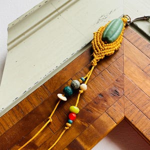May include: A handcrafted macramé pendant necklace featuring a green stone and colourful beads. The pendant is made of yellow macramé and has a green, oval-shaped stone. The necklace has a string of colourful beads in various shapes and colours.