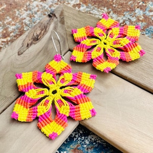 May include: A pair of handmade flower-shaped earrings. The earrings are crafted with macrame knots in vibrant colors: yellow, pink, and orange. The earrings are displayed on a wooden surface.