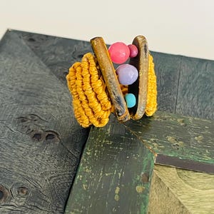 May include: A distinctive ring with a woven yellow band, two dark brown wooden bars, and colourful beads. The beads are pink, lavender, and turquoise. The ring is set against a textured, dark green and grey background.