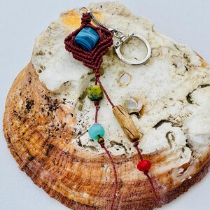 May include: A handcrafted macramé keyring with a dark red woven square holding a blue stone. Below, a string of beads in green, turquoise, and red, plus a wooden bead, hangs from the macramé. A silver keyring is attached.