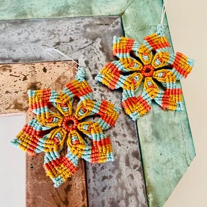 May include: A pair of handmade macrame flower earrings. The earrings feature a vibrant color scheme of orange, yellow, and light blue. The floral design is intricate, with each petal meticulously crafted. The earrings hang from silver-colored hooks.