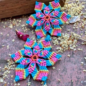 May include: Two handmade macrame flower earrings. Each earring features a vibrant rainbow color scheme with pink, blue, orange, and green. The earrings are made with knotted cord and have a central circular design.