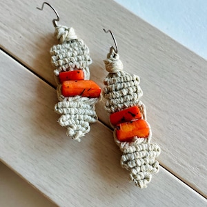 May include: A pair of handmade macrame earrings. Each earring features a beige woven design with three orange-red beads. The earrings have silver-colored ear wires and are displayed on a light wood surface.