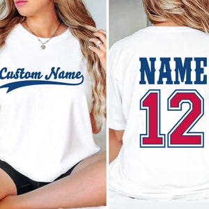 May include: White t-shirt with a blue and red baseball jersey design. The front of the shirt says "Custom Name" in a blue cursive font with a blue and white baseball-style stripe. The back of the shirt says "NAME 12" in a blue and red font with a blue and white baseball-style stripe.