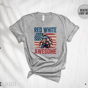 May include: A heather light grey t-shirt featuring a graphic of a superhero holding a shield in front of a distressed American flag. The text RED WHITE is printed in blue above the figure, and the word AWESOME is printed in red below the figure.