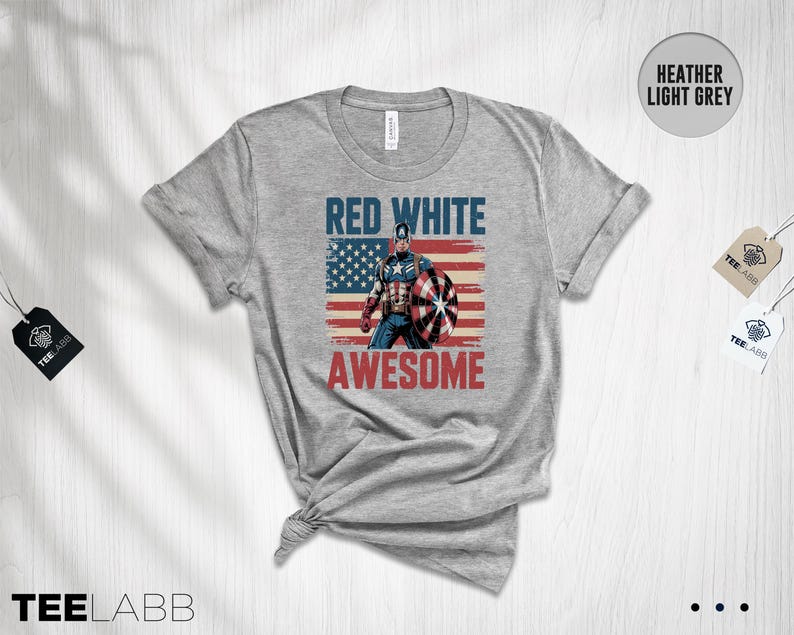 May include: A heather light grey t-shirt featuring a graphic of a superhero holding a shield in front of a distressed American flag. The text RED WHITE is printed in blue above the figure, and the word AWESOME is printed in red below the figure.